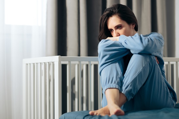 Woman in blue pajamas sitting on a bed with her knees pulled to her chest, looking pensive and distressed, in a room with a crib in the background.