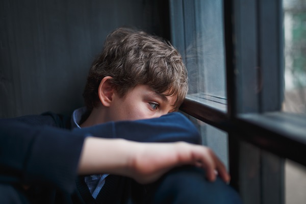 A teenager sits by a window with arms folded, feeling depressed.