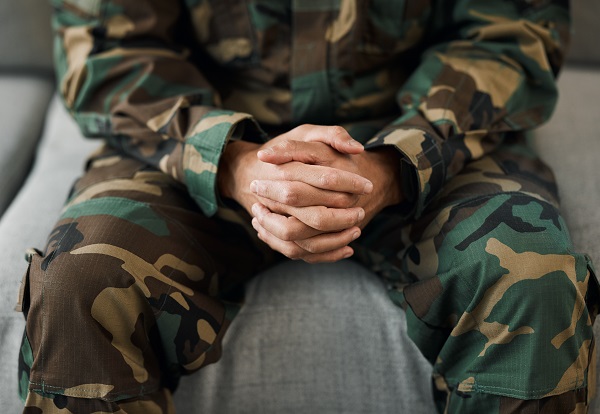 A person dressed in military camouflage sits with their hands clasped, conveying a sense of contemplation or seeking support. 